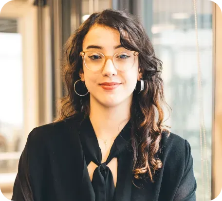 Mulher jovem de cabelos cacheados, usando óculos e brincos de argola, em ambiente interno.